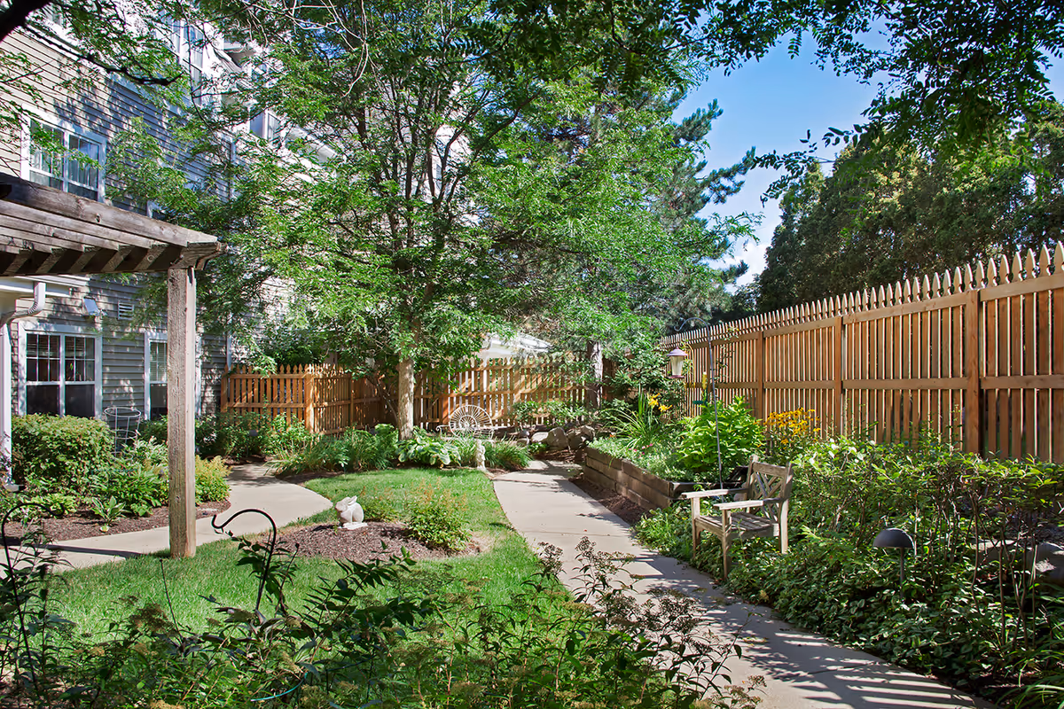 A sunny outdoor garden area with a paved walkway, green grass, various plants, and flowers. There is a wooden fence on the right side and a multi-story building on the left. A wooden pergola is partially visible on the left, and there are benches and garden decorations along the path.