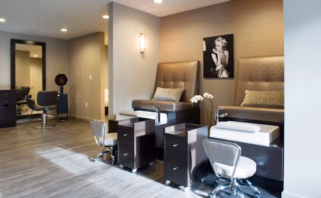 Interior view of a modern salon area in a senior living facility with two pedicure stations featuring cushioned seats and footrests, two rolling chairs, a hair styling chair, and a large mirror on the wall. A black and white portrait of Marilyn Monroe hangs on the wall above the pedicure stations.