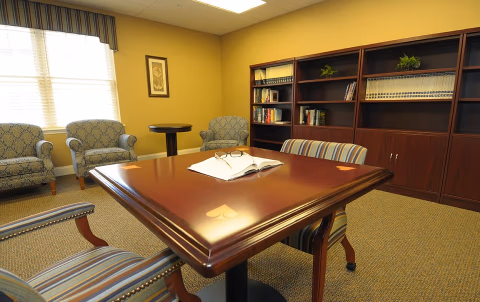 A cozy room with a large wooden table featuring heart-shaped inlays, surrounded by striped and patterned upholstered chairs. In the background, there are three patterned armchairs near a window with blinds and a valance. A tall wooden bookshelf filled with books and decorative plants lines the wall.