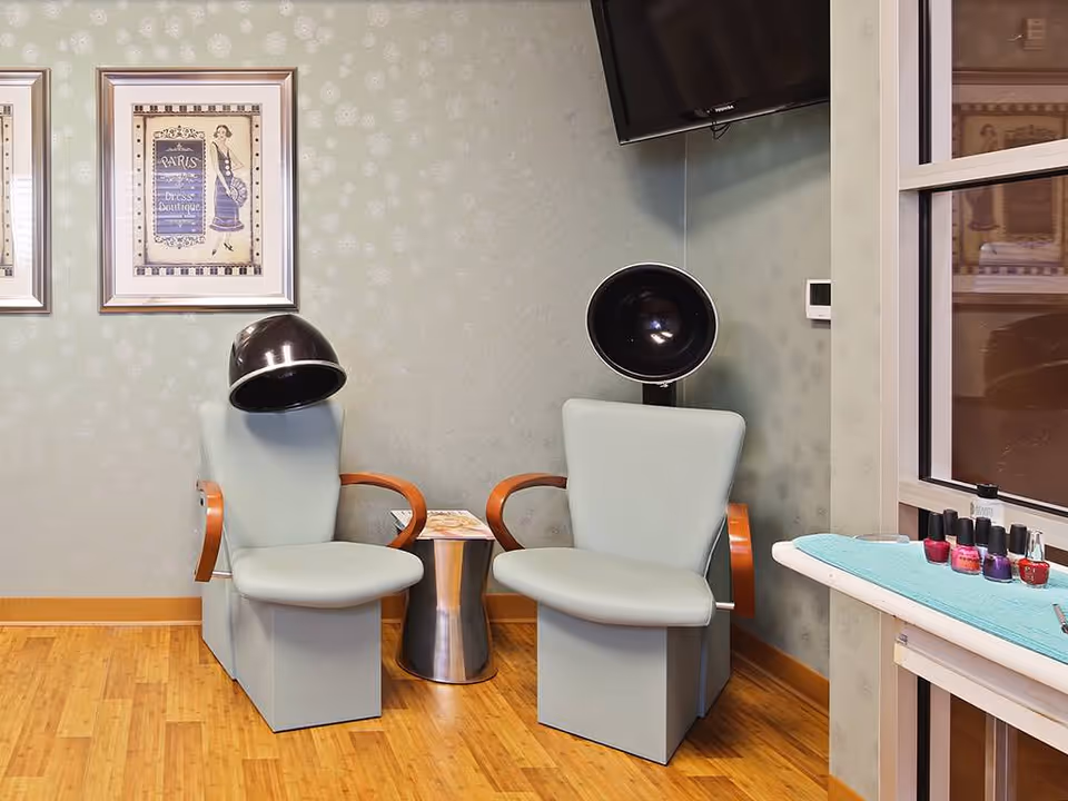 A salon area with two light blue salon chairs equipped with black hair dryers. There is a small metallic side table between the chairs. On the right side, a counter holds several bottles of nail polish. The walls are light green with a subtle pattern, and two framed vintage-style pictures hang on the wall.