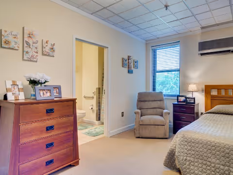 A cozy bedroom in a senior living facility with a wooden dresser holding framed photos and a vase of flowers, a comfortable recliner chair near a window with blinds, a nightstand with a lamp and photo frames, and a bed with a quilted bedspread. The room has beige walls and carpet, and an open door reveals a bathroom with a toilet and grab bars.