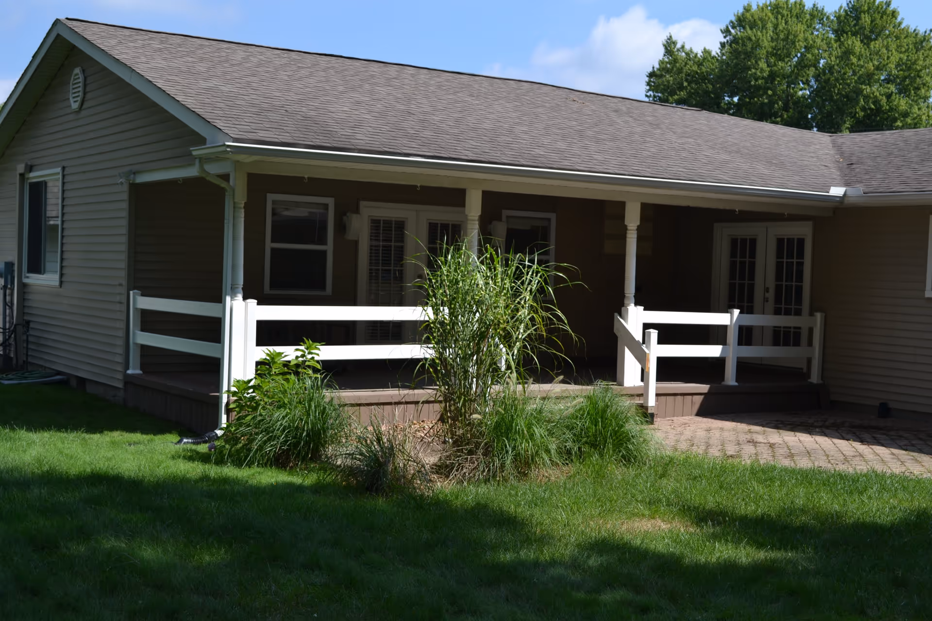 Exterior view of a single-story building with beige siding and a dark shingled roof. The building has a covered porch with white railings and columns. There are two sets of glass doors and a window visible on the porch. In front of the porch is a small garden area with tall green plants and grass surrounding it. Trees and a blue sky with some clouds are visible in the background.