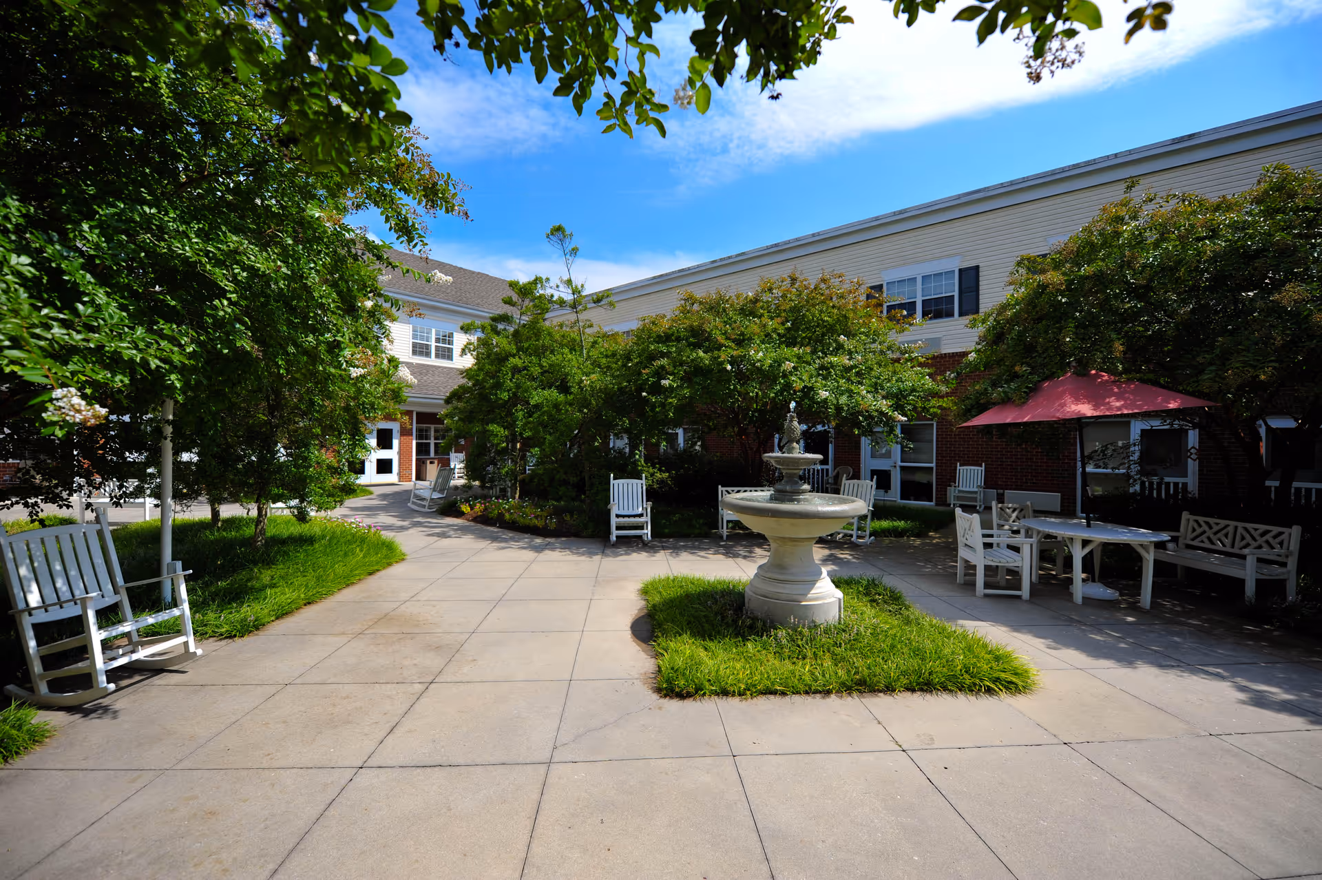 Outdoor courtyard area at Morningside in the West End featuring a central stone fountain surrounded by green grass and trees. White rocking chairs and benches are placed along the paved walkways, with a table and chairs under a red umbrella on the right side. The building exterior is visible in the background under a blue sky with some clouds.