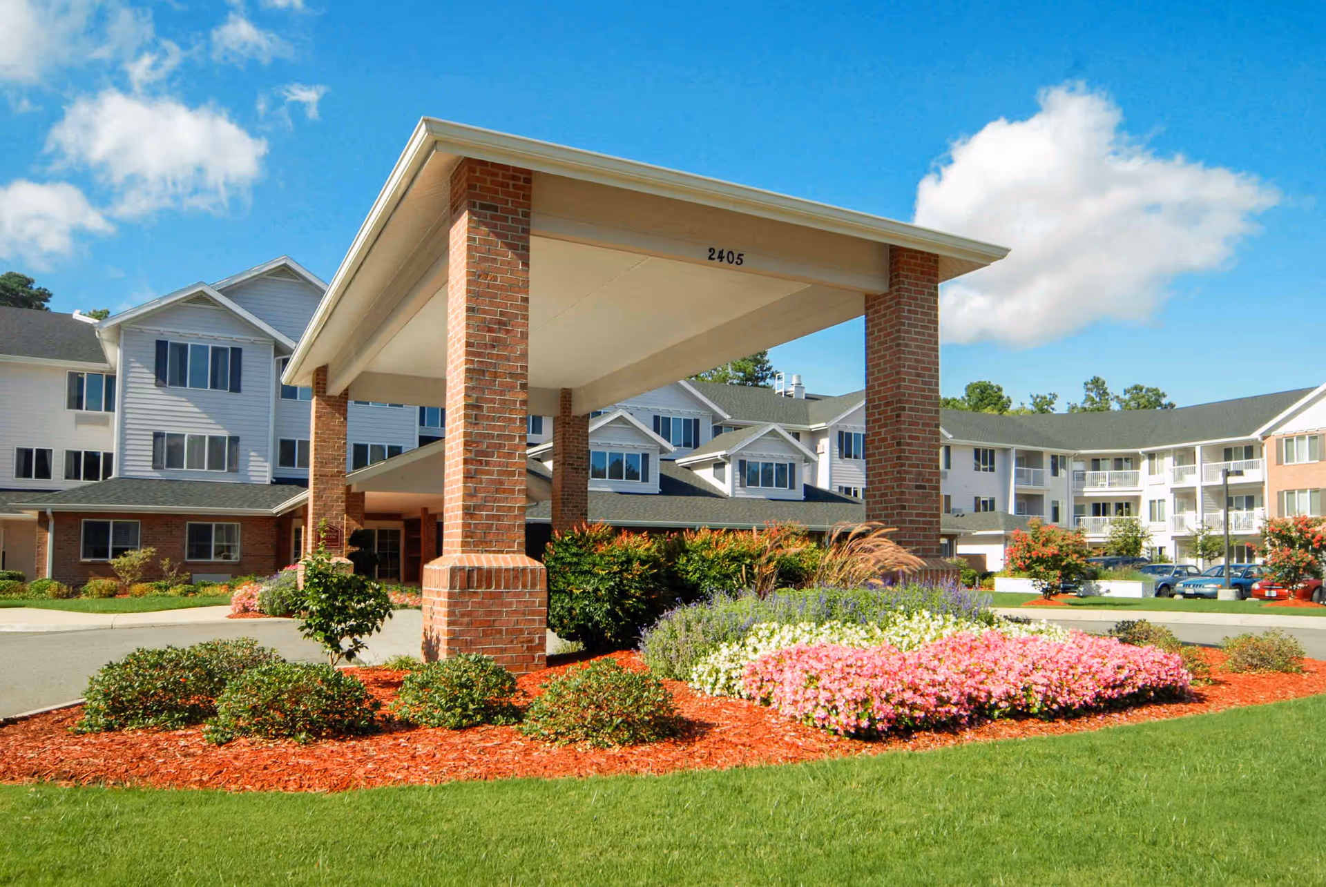 Exterior view of Colonial Harbor Gracious Retirement Living facility showing a covered entrance supported by brick pillars, landscaped flower beds with pink, white, and purple flowers, green bushes, and a well-maintained lawn under a blue sky with some clouds.