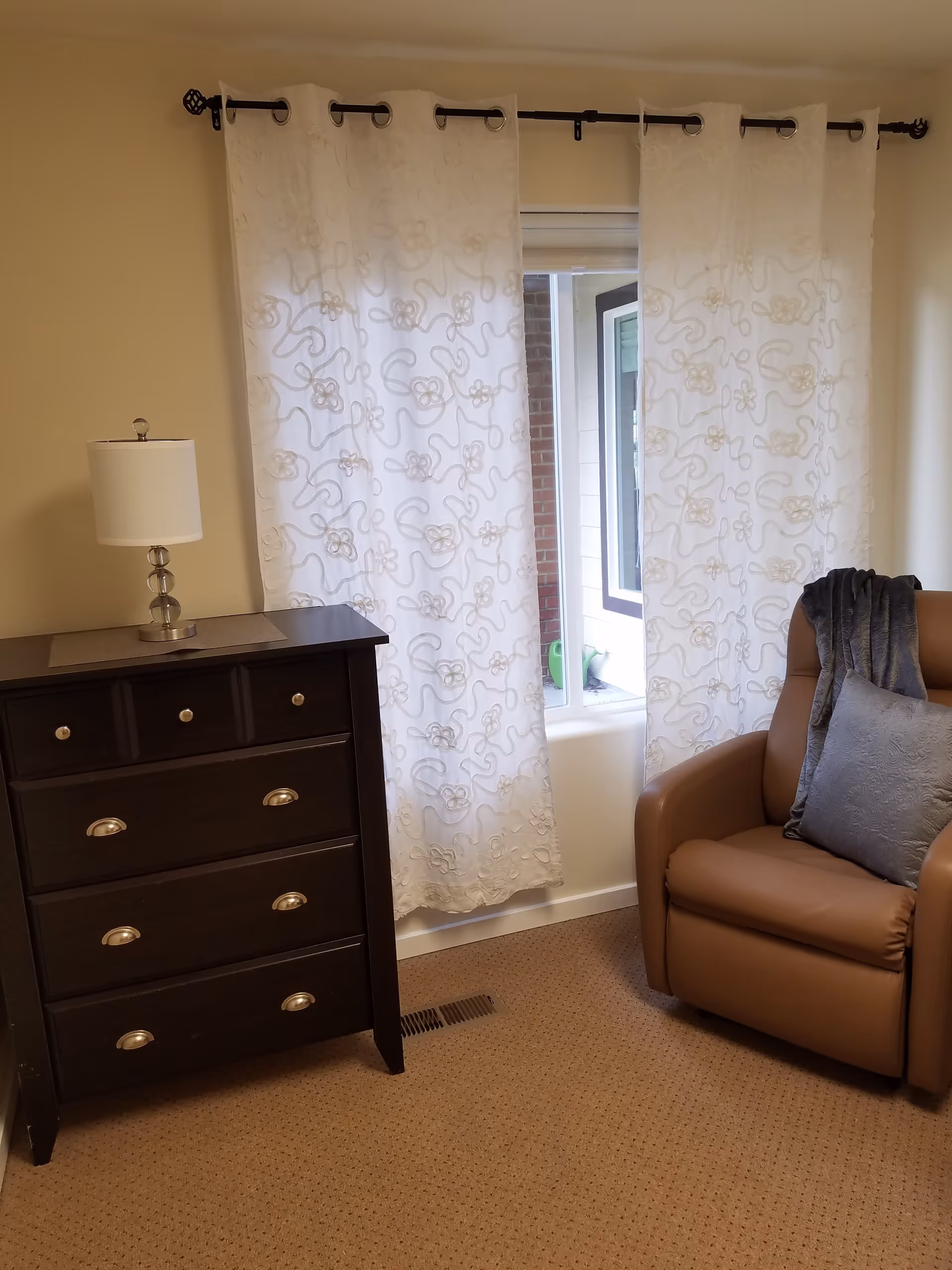A cozy corner of a room featuring a brown cushioned armchair with a gray pillow and a matching throw draped over the back. Next to the chair is a dark wooden dresser with four drawers and a white table lamp on top. Behind the furniture is a window with sheer white curtains that have a floral and swirl pattern, allowing some natural light to filter through.