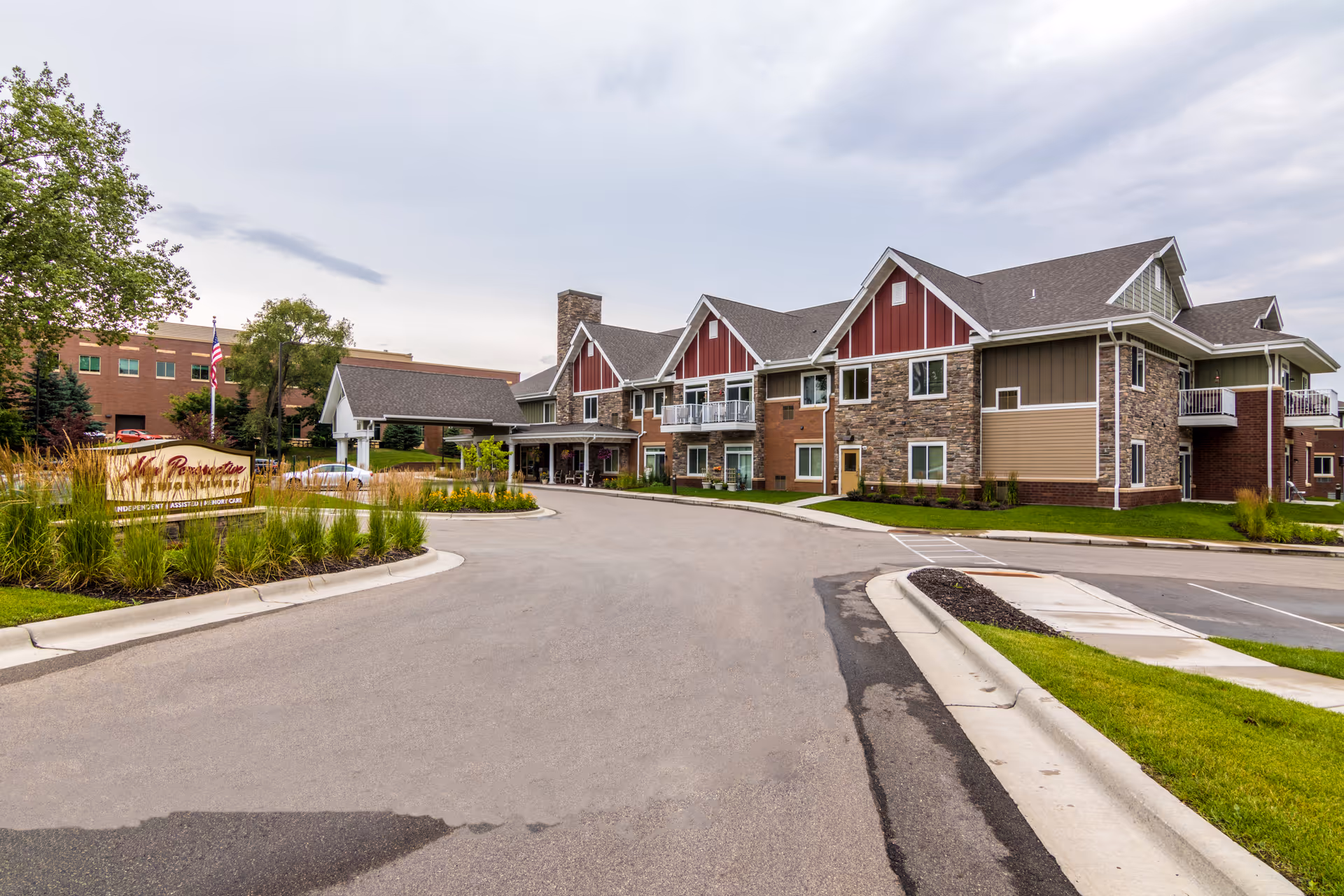 Exterior view of New Perspective Senior Living | Woodbury facility showing a large two-story building with a mix of stone, red siding, and beige panels. The building has multiple windows and balconies. There is a driveway leading to a covered entrance with landscaping and a sign on the left side. The sky is cloudy.