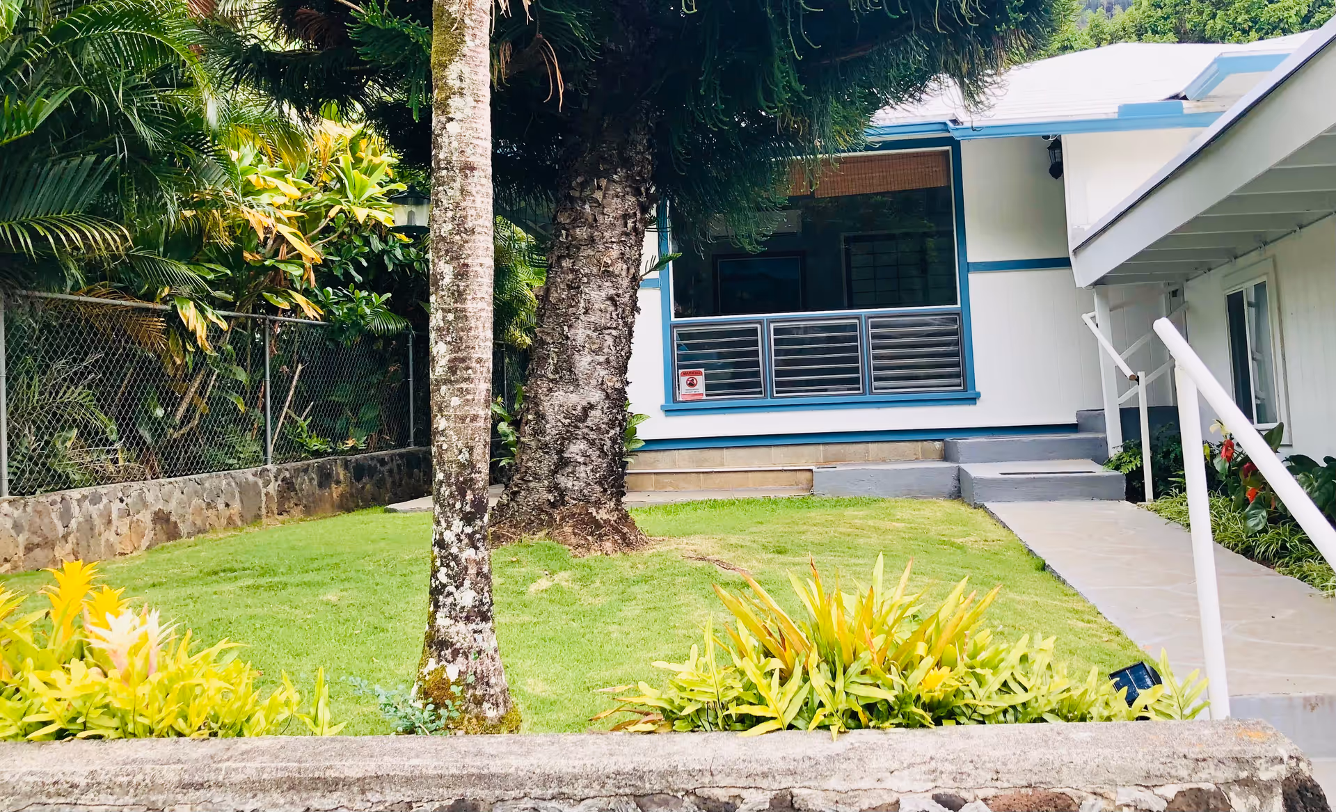 View of a single-story building with a small porch and steps leading up to it, surrounded by green grass, tropical plants, and trees. A concrete pathway with a white handrail runs alongside the building. The area is enclosed by a chain-link fence with dense foliage behind it.