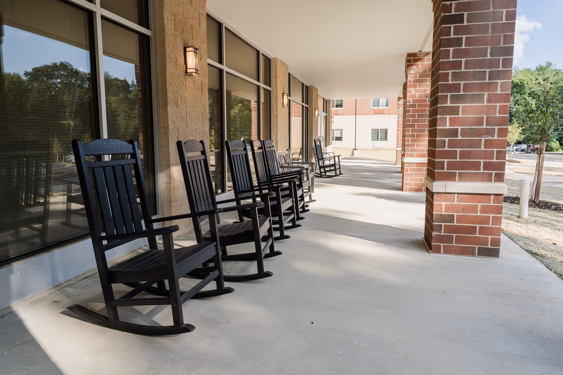 A row of black wooden rocking chairs lined up on a covered outdoor patio with brick columns and large windows reflecting trees and sky.