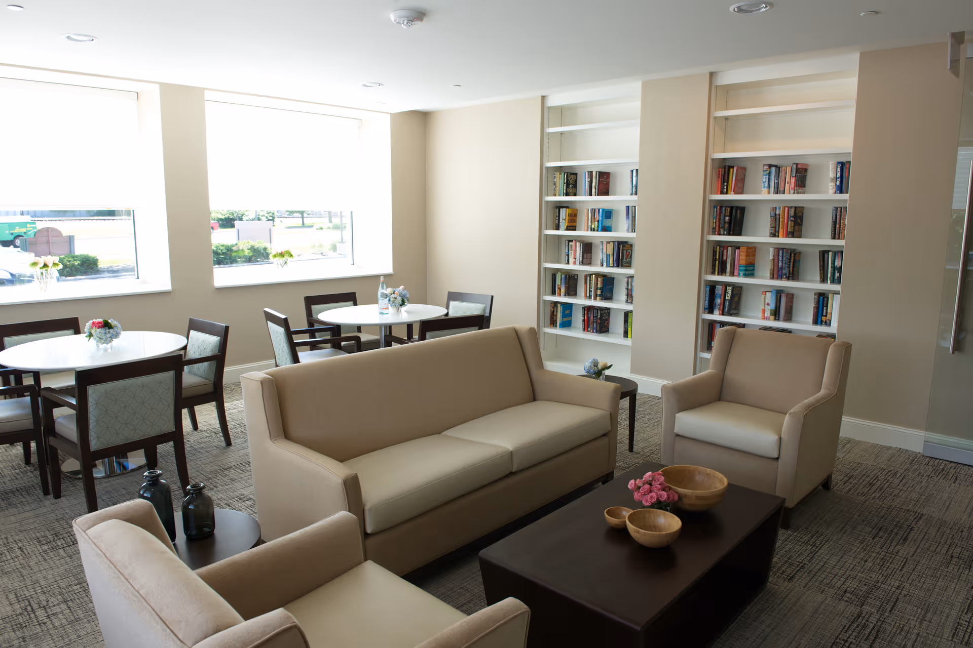 A bright and cozy common area with beige sofas and armchairs arranged around a dark wooden coffee table. The room features built-in white bookshelves filled with books, round tables with chairs near large windows letting in natural light, and small floral decorations on the tables.
