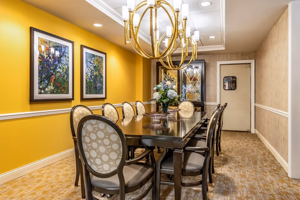 A dining room with a long dark wooden table surrounded by eight upholstered chairs with circular patterned backs. The walls are painted yellow on one side with two colorful framed paintings, and beige on the other side. A large gold chandelier with multiple lights hangs above the table. At the far end of the room, there is a glass-fronted cabinet with decorative items and a closed white door.