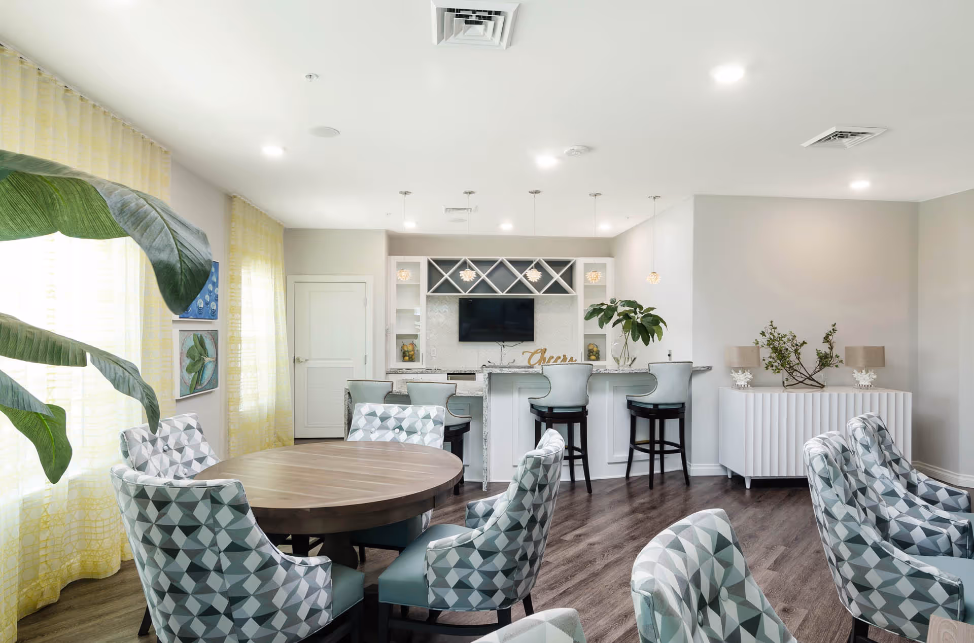 A bright and modern common area with a round wooden table surrounded by patterned upholstered chairs. In the background, there is a white bar counter with four light blue bar stools, a mounted flat-screen TV, and decorative shelves. The room has large windows with yellow curtains, wooden flooring, and various plants and decor items.