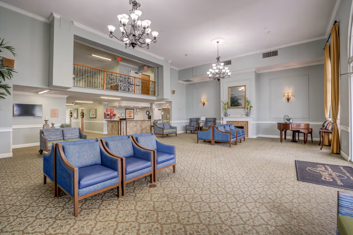 Spacious senior living lobby with multiple blue chairs, chandeliers, a reception desk, piano, and an upper balcony.