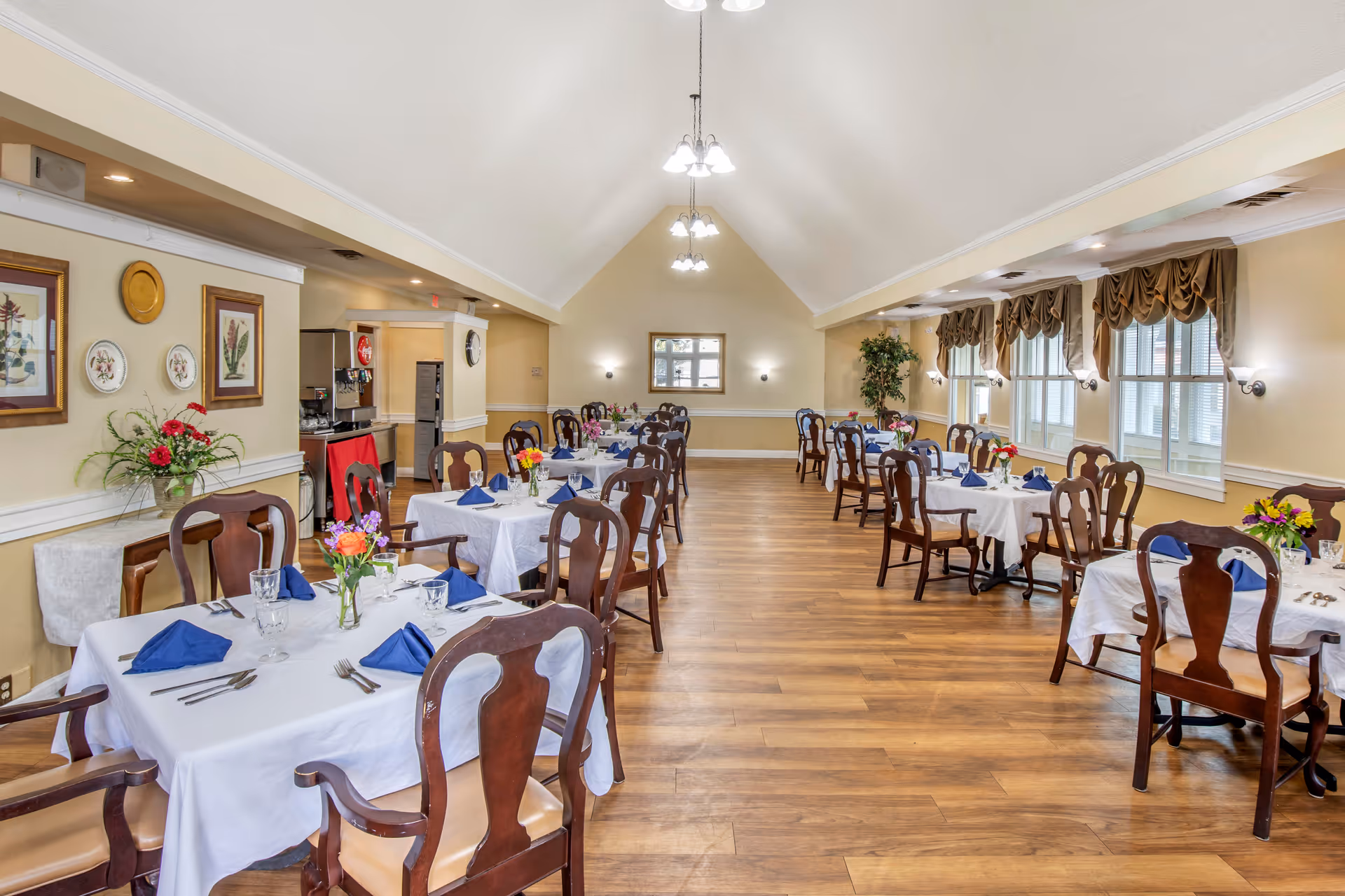 A spacious dining room with wooden floors and multiple tables covered with white tablecloths, each set with blue folded napkins, glassware, and silverware. The room has high vaulted ceilings with hanging light fixtures, large windows with brown drapes on the right side, and framed botanical artwork on the left wall. There are floral centerpieces on each table and a beverage station in the back left corner.
