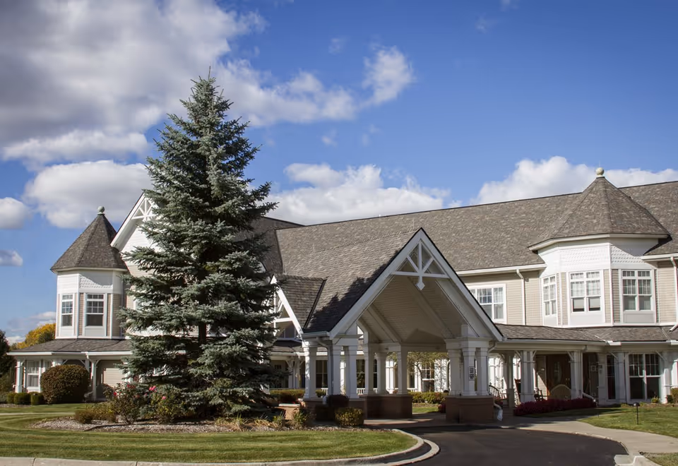 Front exterior of a large two-story senior living building with a porte-cochere, landscaped lawn, and a tall evergreen tree under a partly cloudy sky.