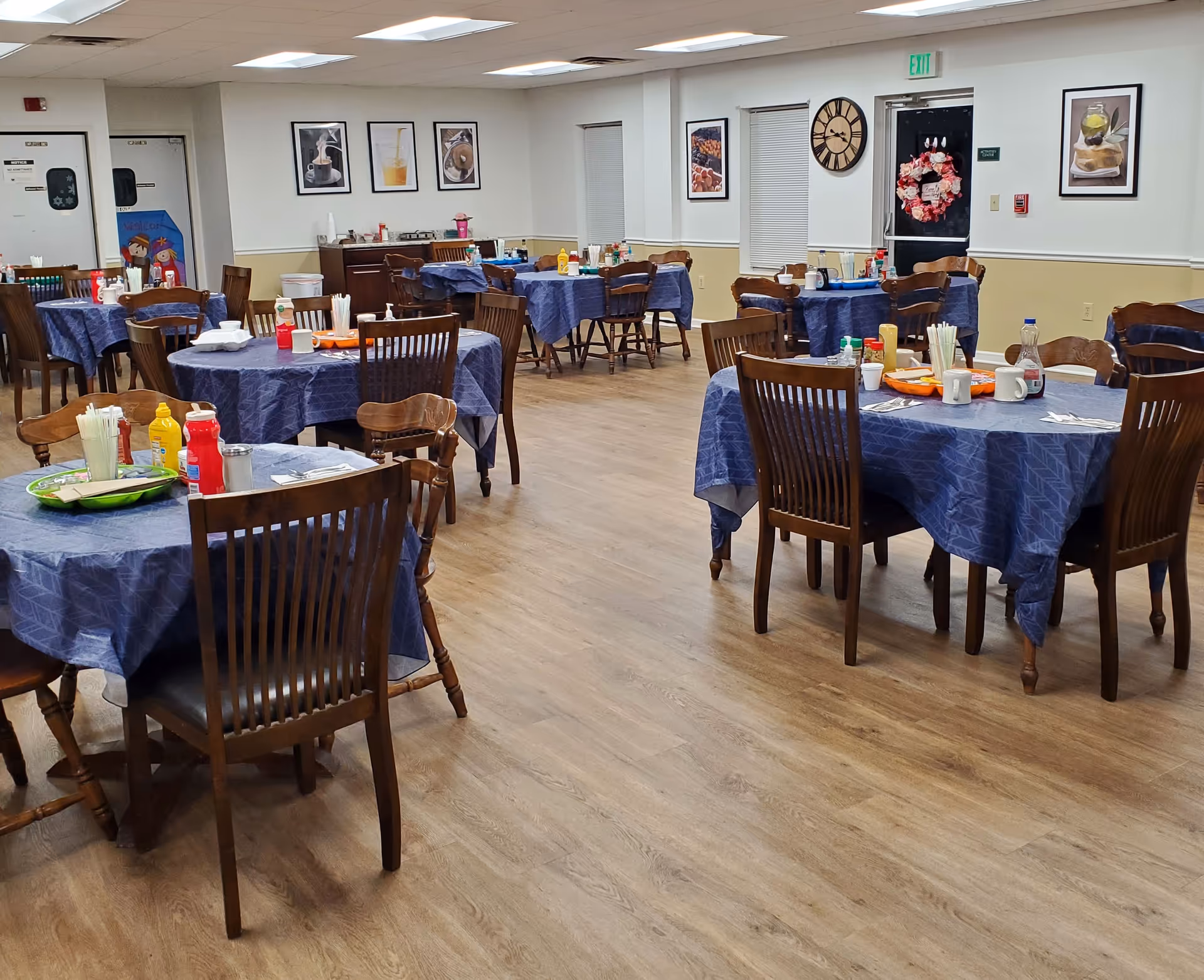 A dining room in Pleasant Villa Retirement Home with several round tables covered with blue tablecloths. Each table has wooden chairs around it and is set with condiments, napkins, and utensils. The room has light-colored walls with framed pictures and a large clock. The floor is wood, and there is a door with a wreath and an exit sign above it.