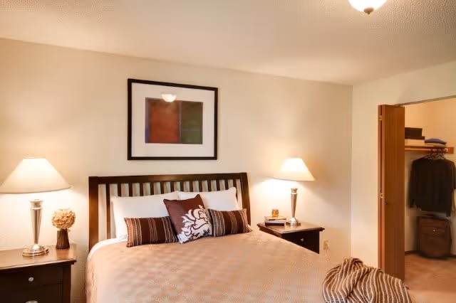 A cozy bedroom with a neatly made bed featuring a beige bedspread and decorative pillows. There are two bedside tables with lamps on each side of the bed. A framed abstract artwork hangs above the headboard. An open closet with clothes and storage boxes is visible on the right side of the room.