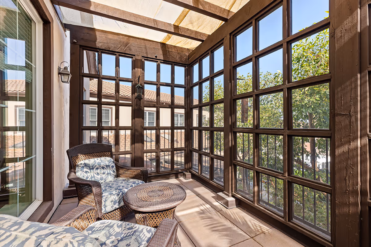 A sunlit enclosed balcony or patio area with large grid-style windows and a translucent roof. The space features two wicker armchairs with patterned cushions and a matching round wicker table. Outside the windows, trees and rooftops are visible under a clear blue sky.