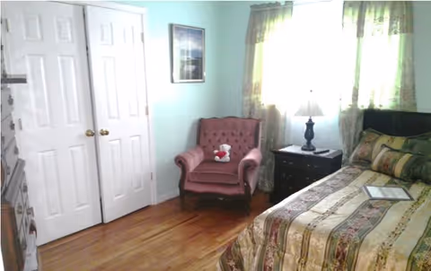 A cozy bedroom with a patterned bed, a pink upholstered armchair holding a teddy bear, a nightstand with a lamp, closet doors, and a window with green curtains.