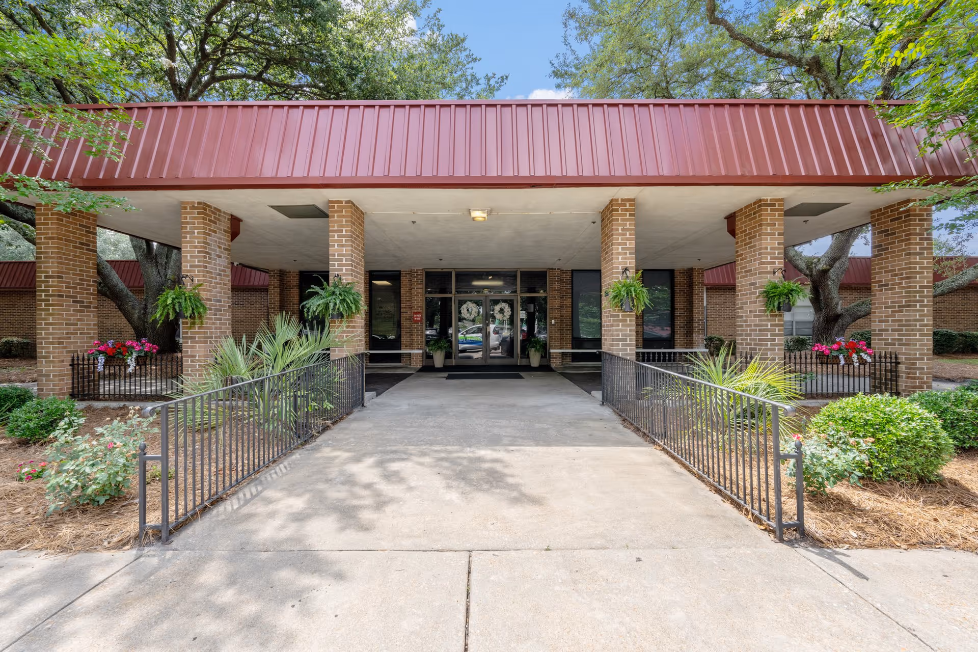 Entrance of Sumter East Health and Rehabilitation Center showing a covered walkway supported by brick columns, with hanging plants and landscaped greenery on either side of the pathway leading to glass doors.
