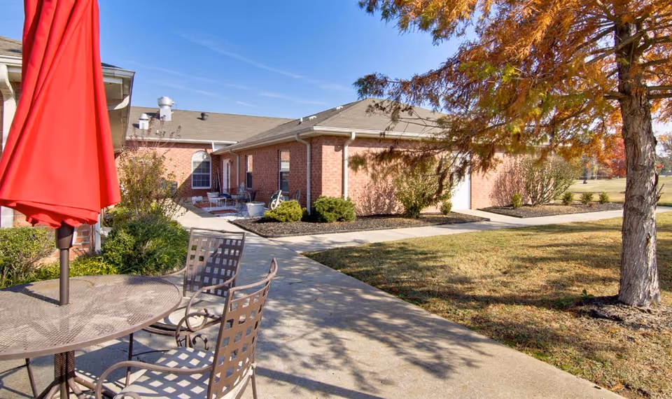 Outdoor courtyard with a patio table and chairs under a red umbrella beside a walkway and brick building with landscaping.