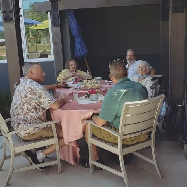 A group of five elderly people sitting around a round table covered with a pink tablecloth outdoors, enjoying snacks and drinks. The setting appears to be a patio area with chairs and an umbrella in the background.