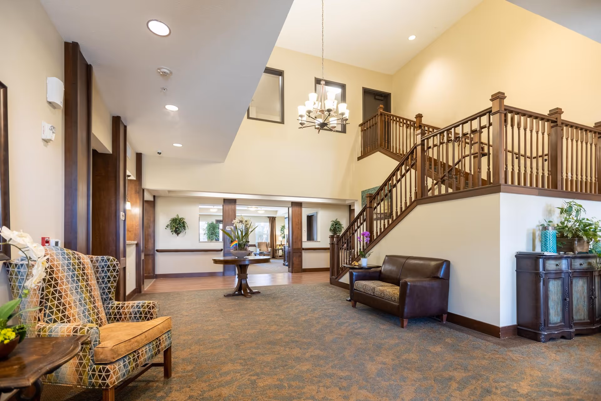 Spacious senior living lobby with seating, a central table, plants, and a wooden staircase to an upper level.