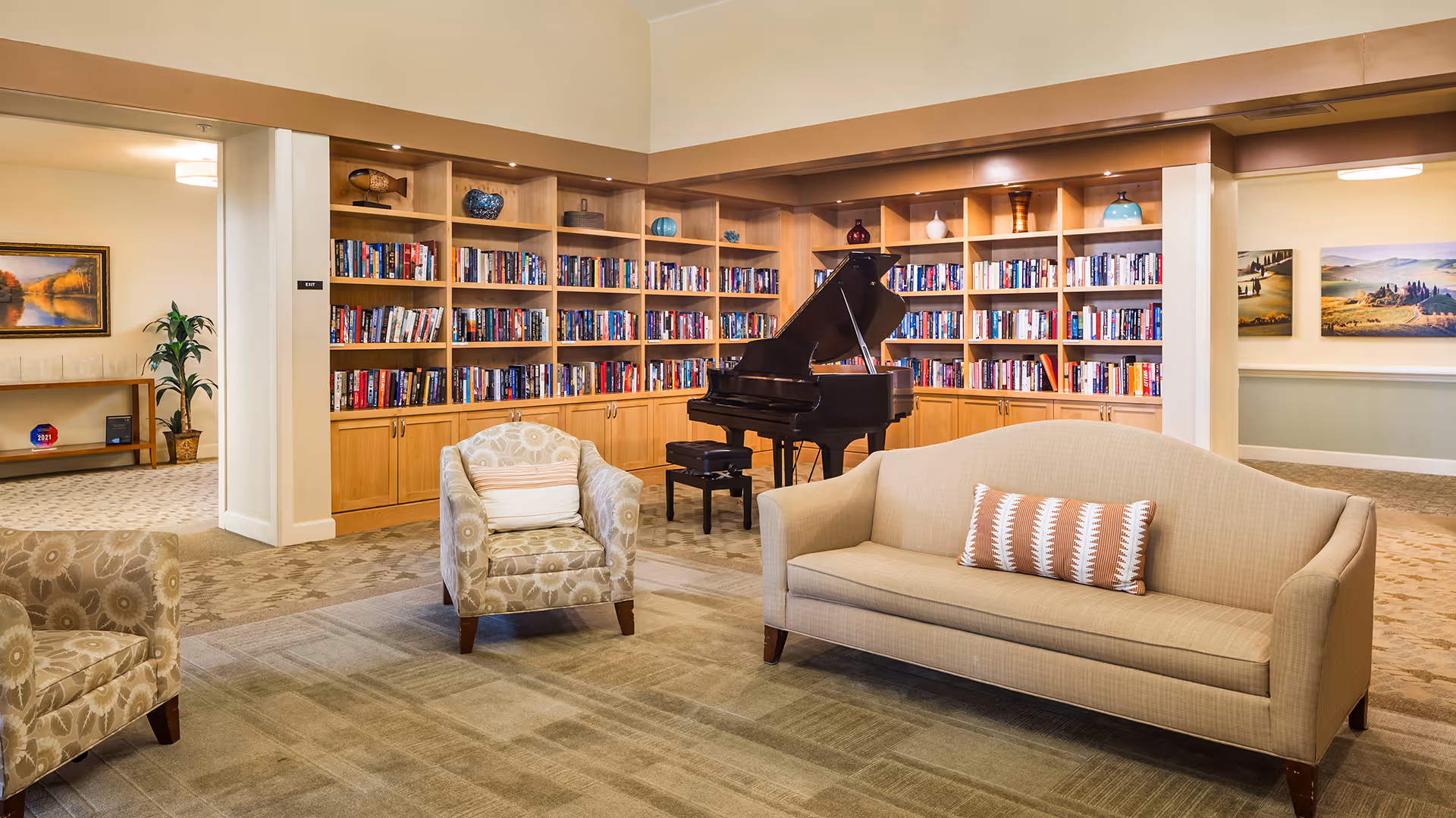 A cozy living room area with a beige couch and two patterned armchairs arranged around a carpeted floor. Behind the seating is a large wooden bookshelf filled with books and decorative items. A black grand piano with a matching bench is positioned in front of the bookshelf. The room has warm lighting and neutral-colored walls, with artwork visible in adjacent rooms.