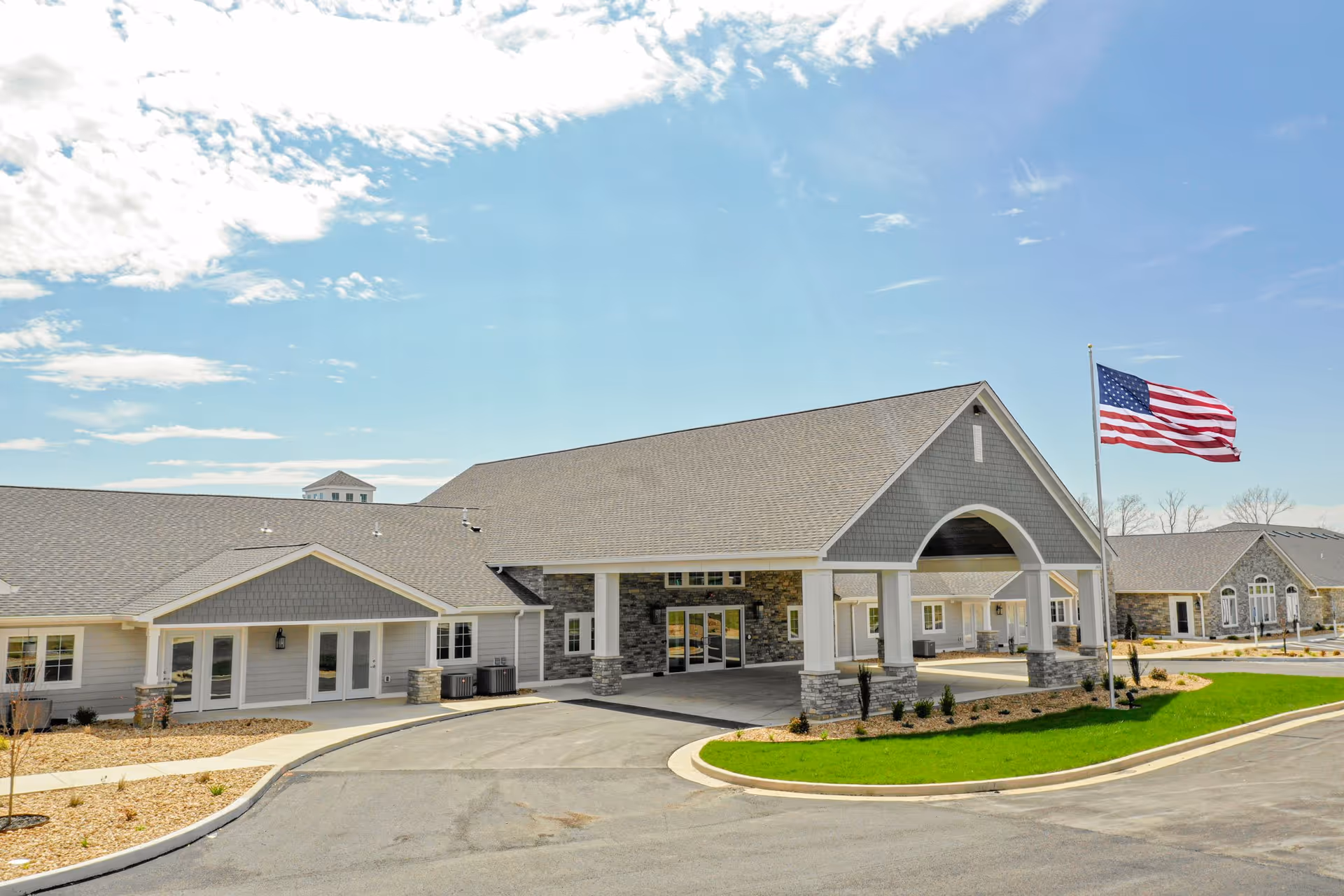 Exterior view of Cedarhurst Senior Living of West Plains building with a large covered entrance, stone and gray siding facade, an American flag on a flagpole, and a clear blue sky with some clouds.