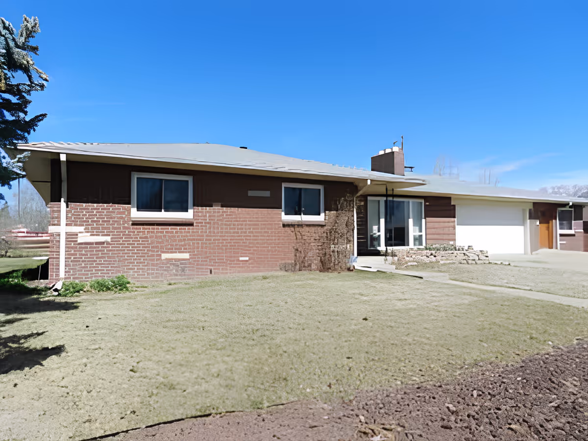 Single-story brick and wood residential building with a sloped roof, two windows, a glass front door, and an attached garage. The building is surrounded by a grassy lawn and a clear blue sky is visible above.