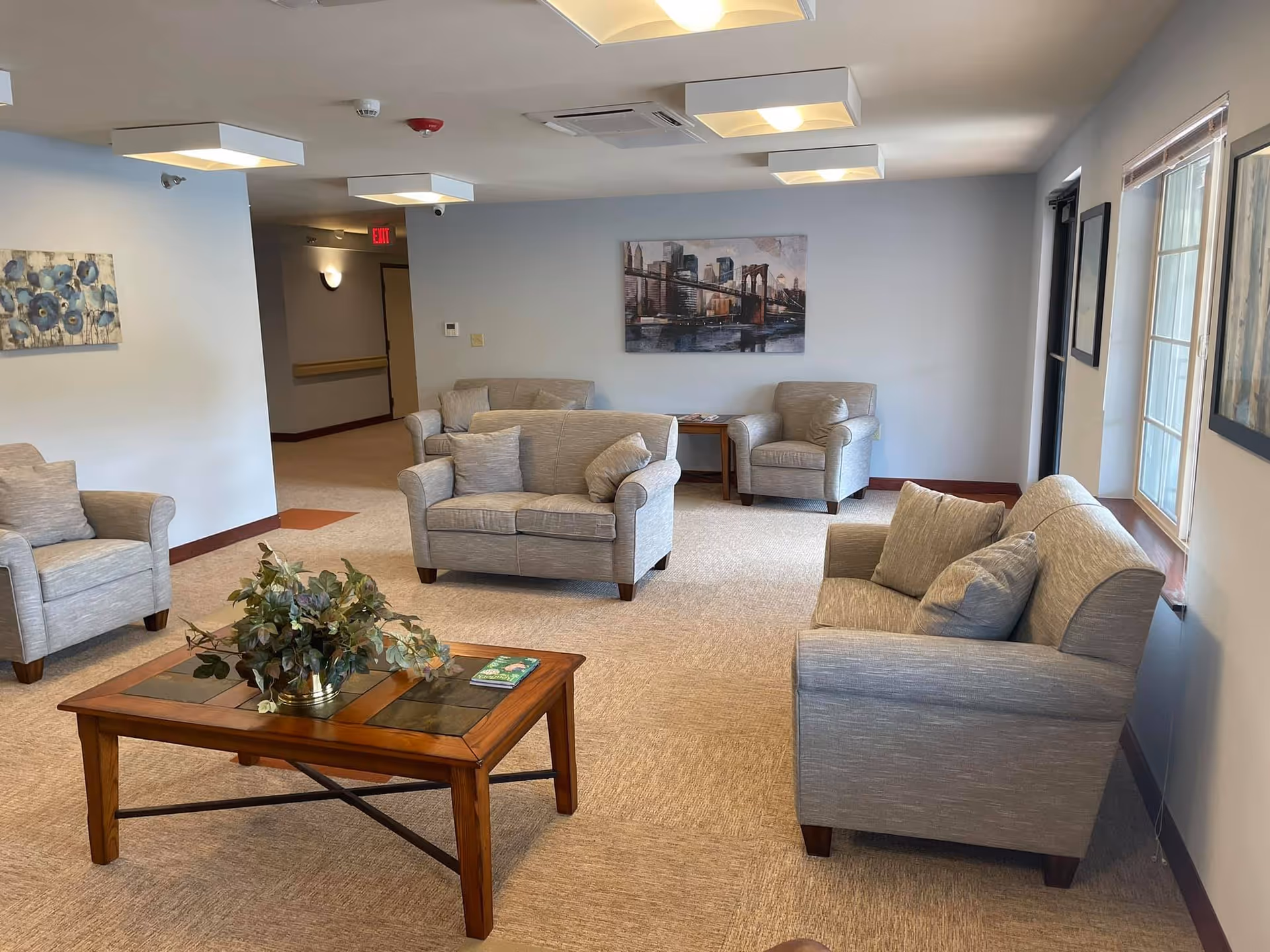 Bright common sitting area with multiple gray upholstered chairs and sofas arranged around a wooden coffee table and windows.