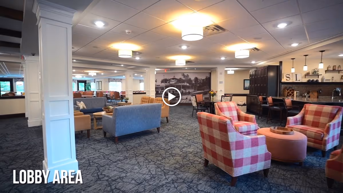 A spacious and well-lit lobby area with multiple seating arrangements including blue and beige sofas, and red and white checkered armchairs around a round ottoman. The room has a carpeted floor, white pillars, and ceiling lights. In the background, there is a bar area with stools and shelves, and a large black and white photo on the far wall.