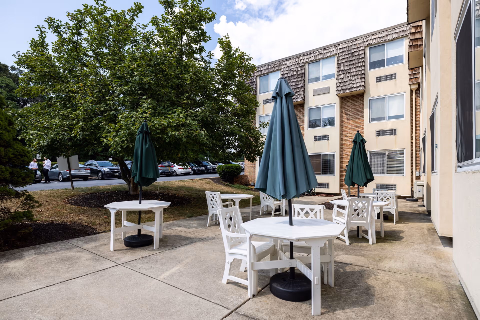 Outdoor patio area with white tables and chairs, each table having a closed green umbrella. The patio is adjacent to a multi-story building with several windows. There are trees and parked cars visible in the background under a partly cloudy sky.