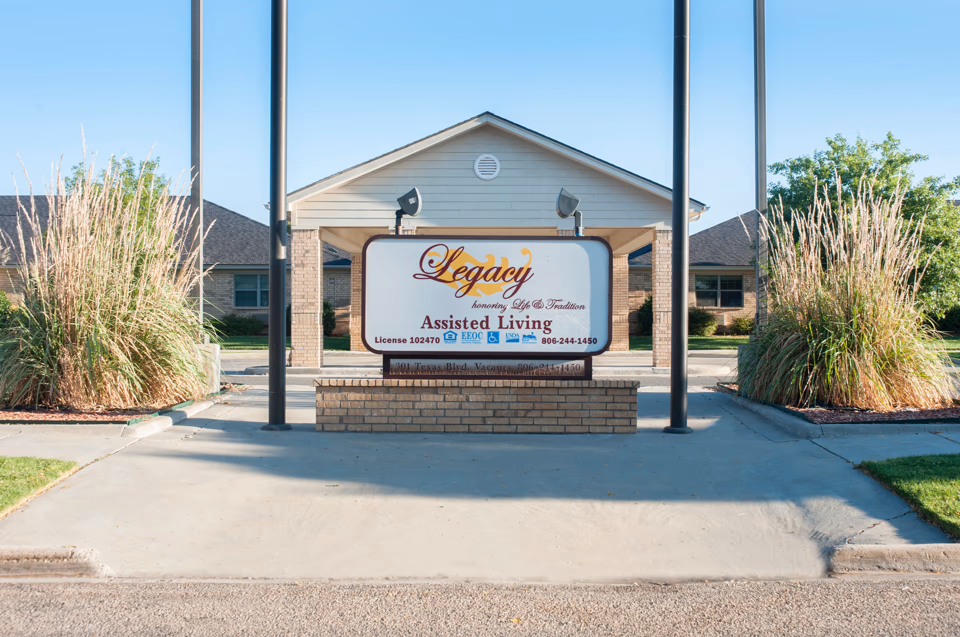 Outdoor view of the entrance sign for Legacy Assisted Living Community, featuring a large sign with the facility's name and contact information, flanked by tall ornamental grasses and a building in the background under a clear blue sky.