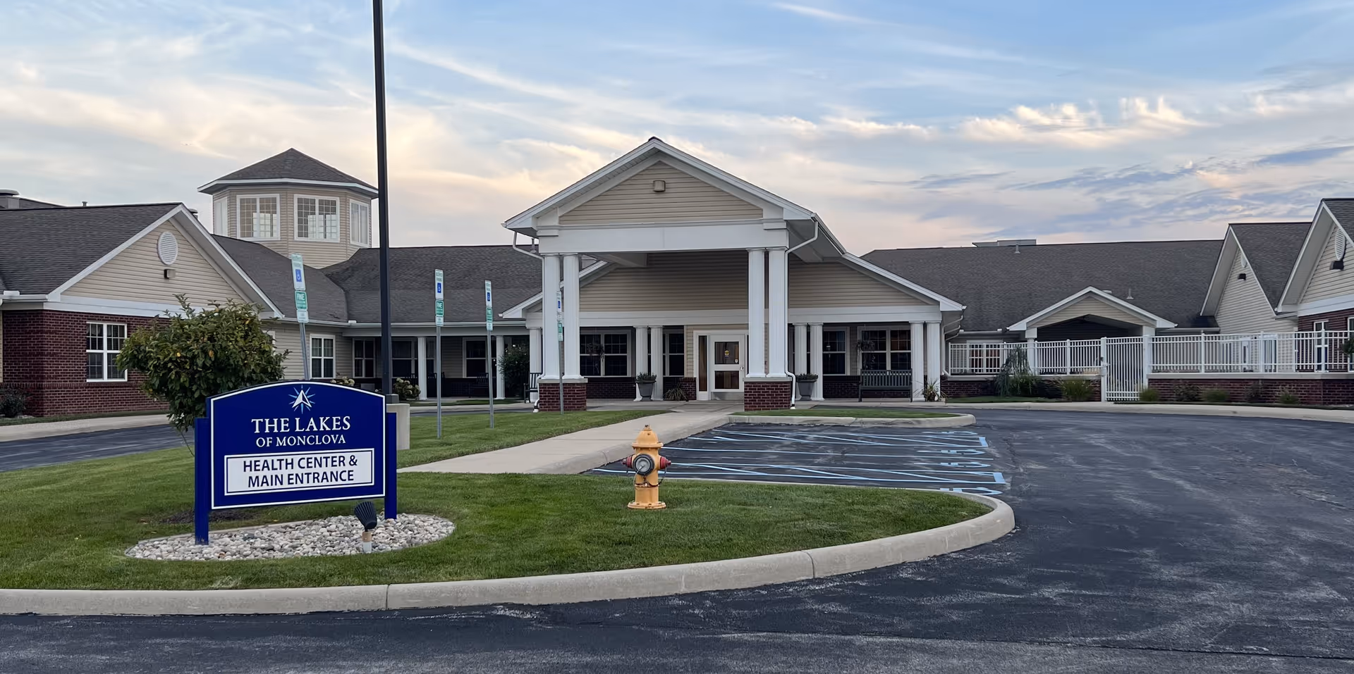 Exterior view of The Lakes of Monclova health center and main entrance building with a parking lot and a blue sign in front on a grassy area. The building has a covered entrance with white columns and a cupola on the roof. The sky is partly cloudy during sunset or sunrise.