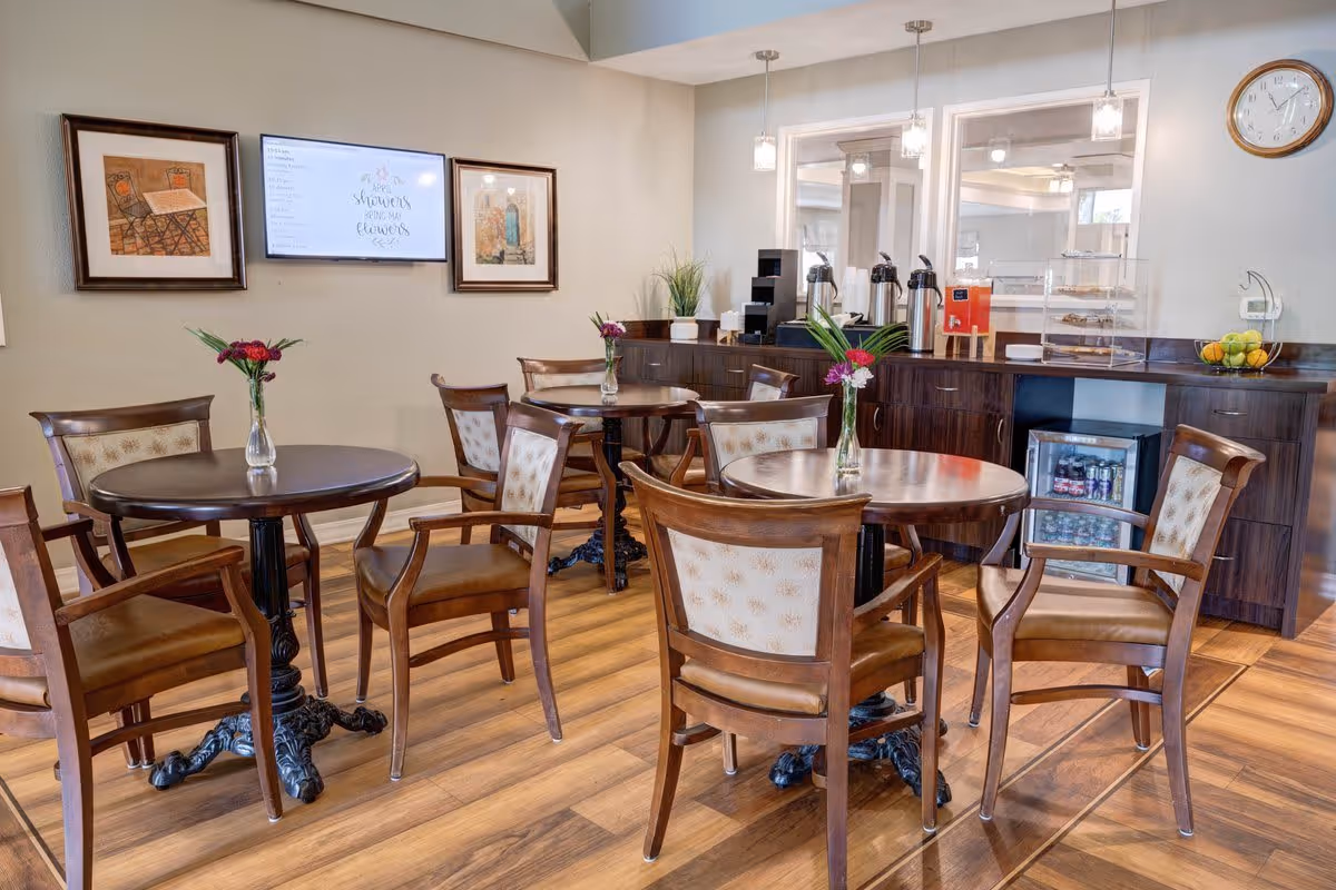 A cozy dining area with round wooden tables and cushioned wooden chairs. Each table has a small vase with flowers. The room features a wooden floor, a counter with coffee dispensers, a small refrigerator stocked with drinks, and a bowl of fruit. On the wall, there are two framed pictures and a digital screen displaying a message. A clock is mounted on the wall above the counter.