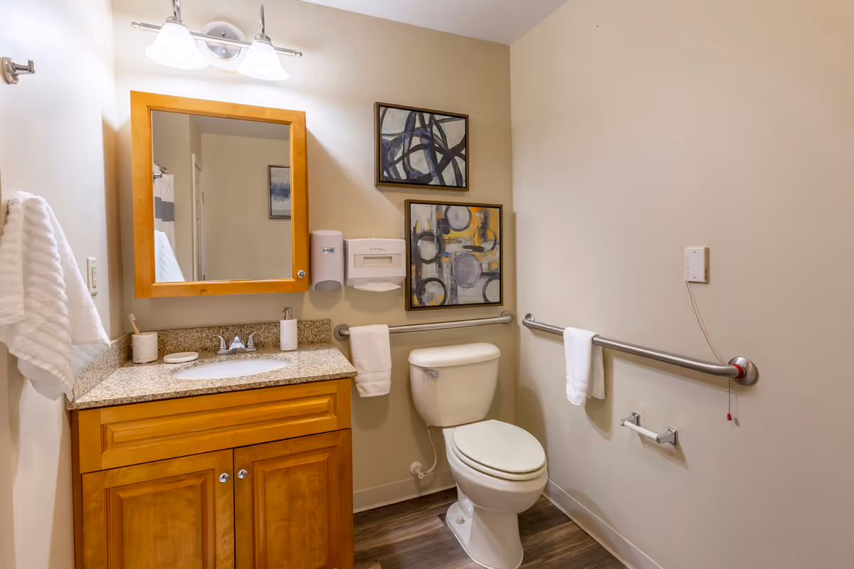 A clean bathroom with a wooden vanity topped with a granite countertop, a sink, and a mirror above it. There are two abstract paintings on the wall above the toilet. The bathroom has grab bars on the walls, a towel rack with white towels, a soap dispenser, and a paper towel dispenser. The floor is wood-style laminate.