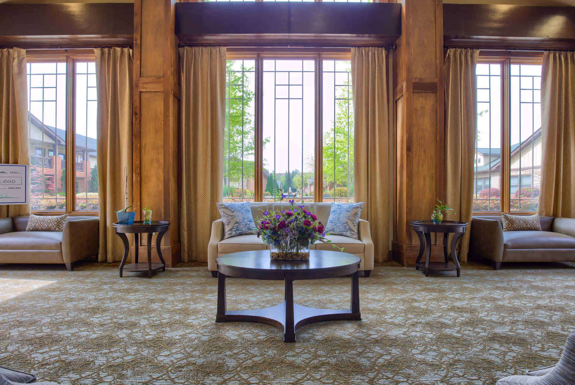Spacious lounge area with large vertical windows, sofas, a round coffee table with a floral arrangement, wood columns and side tables.