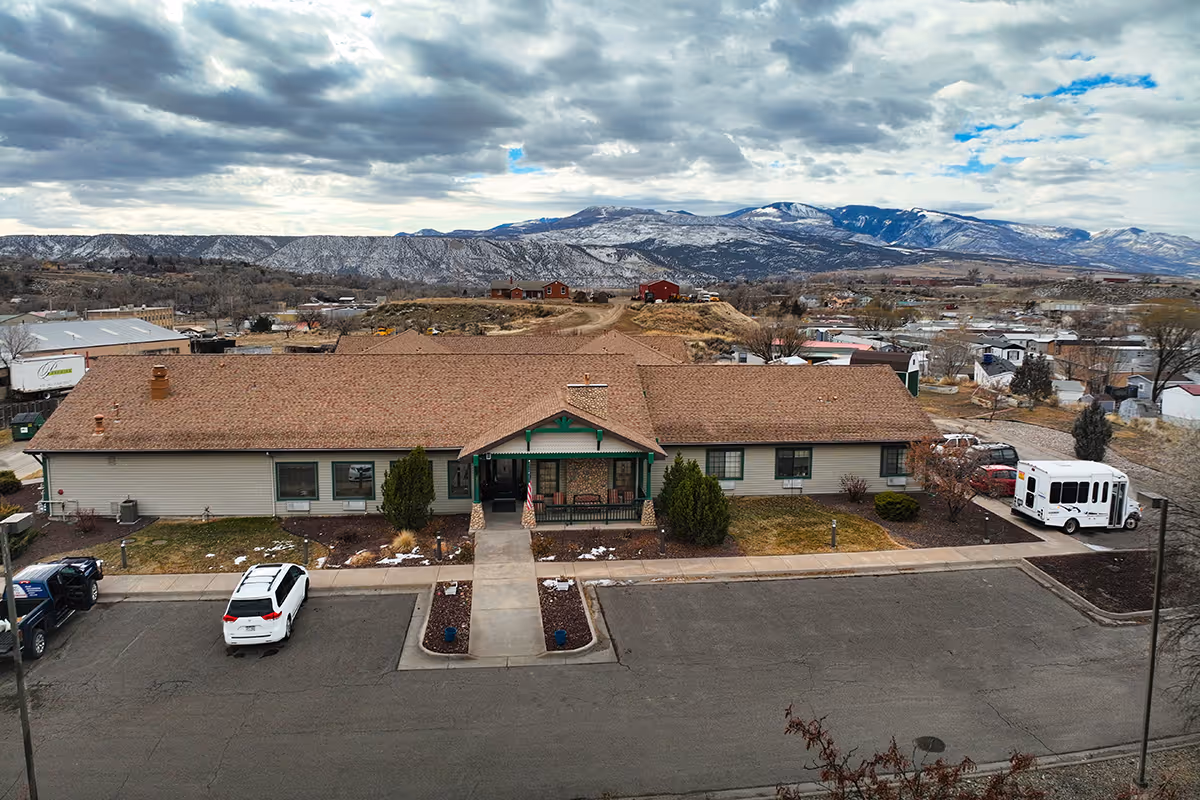 A single-story building with a brown roof and beige siding, identified as Chateau At Rifle, with a parking lot in front containing a few vehicles. The building is set against a backdrop of snow-capped mountains under a cloudy sky.