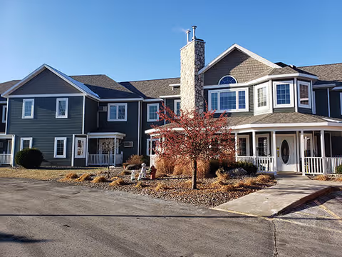 Exterior front view of a two-story assisted living facility building with gray siding, white trim, a stone chimney, and a covered entrance porch. There is a small landscaped area with a tree and bushes in front of the building, and a paved driveway or parking area in the foreground under a clear blue sky.
