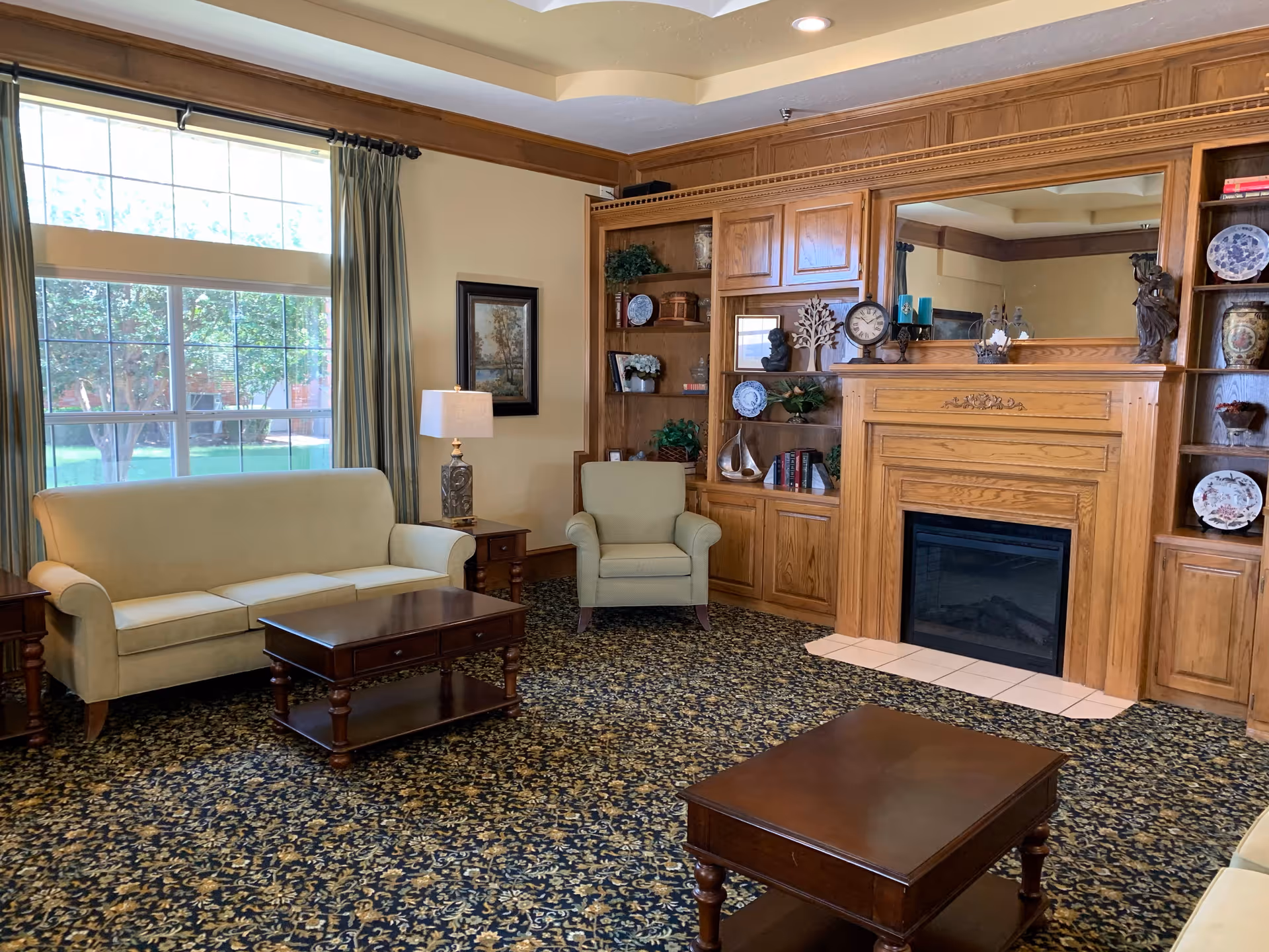 A cozy senior living facility common area with a beige sofa, a matching armchair, two wooden coffee tables, a wooden built-in bookshelf with decorative items, and a fireplace with a large mirror above it. A large window with green curtains lets in natural light, and a table lamp is placed on a side table next to the sofa.