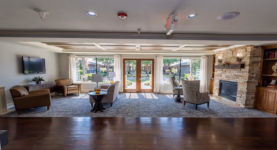 A spacious and well-lit living room area in a senior living facility featuring large windows and glass doors that open to an outdoor garden. The room includes comfortable seating such as armchairs and a sofa, a stone fireplace with a wooden mantel, a wall-mounted TV, and built-in wooden shelves with decorative items. The floor is a combination of dark wood and a large area rug, and the ceiling has recessed lighting and decorative beams.