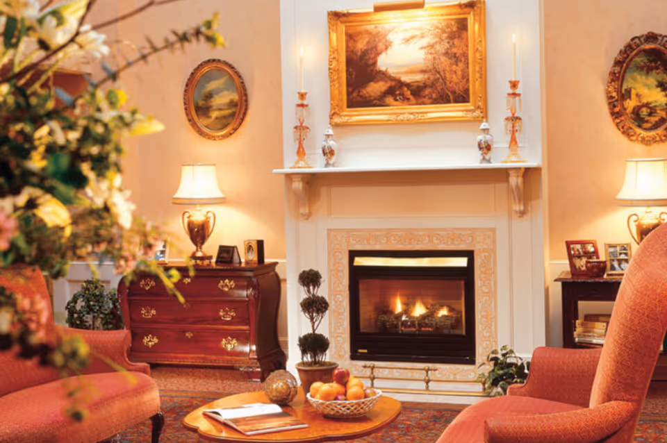 Cozy living room with a lit fireplace, upholstered chairs, a coffee table with a fruit bowl, lamps, and framed artwork.