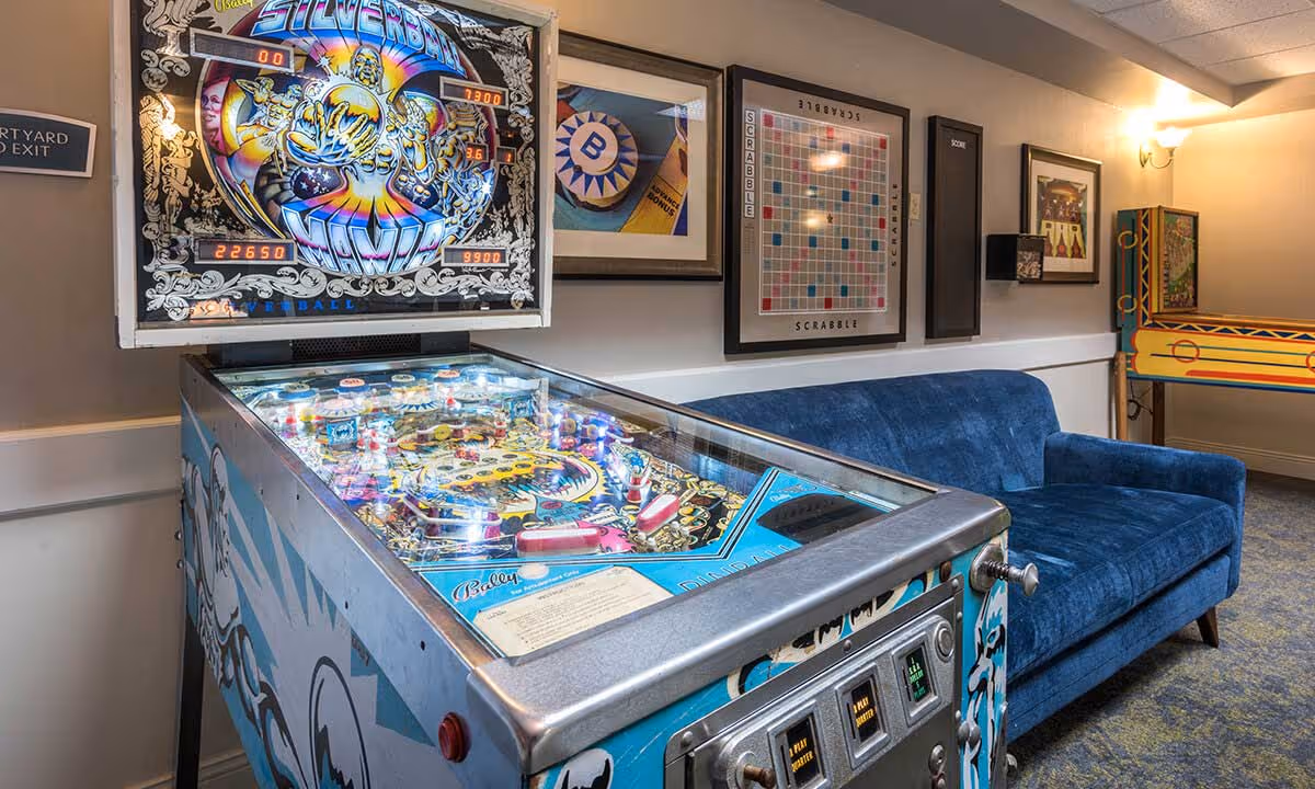 A pinball machine in a lounge area with a blue velvet couch and framed game artwork on the wall.