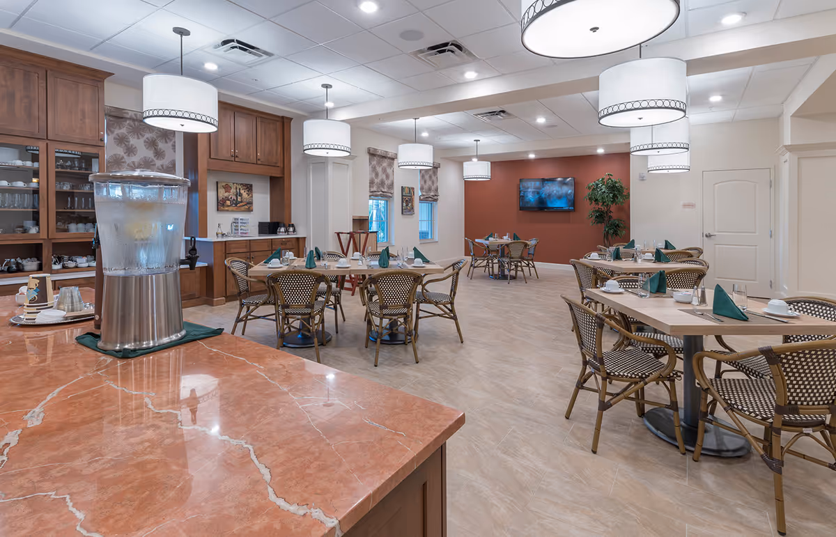 Spacious dining room with multiple set tables and wicker chairs, pendant lights, a TV on the far wall, and a water dispenser on a marble counter.