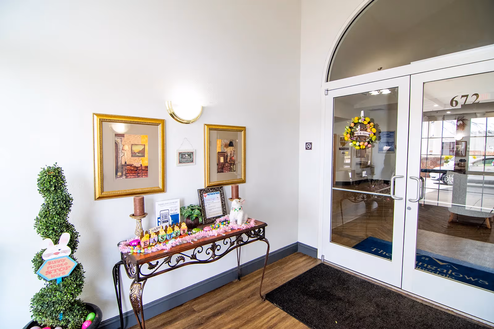 Entrance area of The Bungalows at Springdale facility with glass double doors decorated with a colorful wreath. Inside, a decorative table holds Easter-themed decorations including candles, a small bunny figure, and framed pictures on the wall above. A small potted topiary with a bunny sign stands to the left of the table.