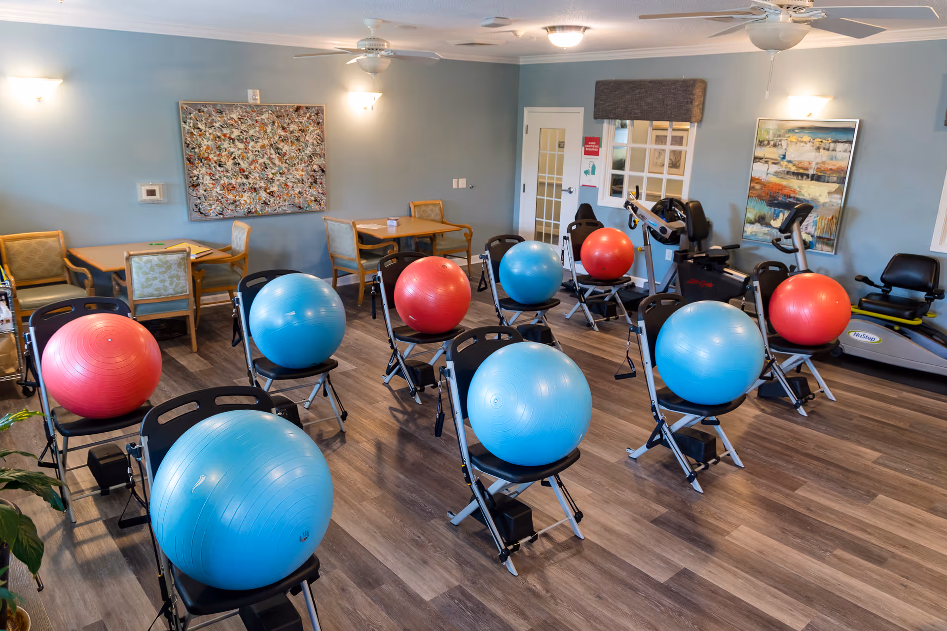 A fitness room with several chairs each holding a large exercise ball, alternating red and blue. The room has wood flooring, light blue walls, ceiling fans, and exercise equipment including recumbent bikes. There are tables and chairs along the back wall and framed artwork on the walls.