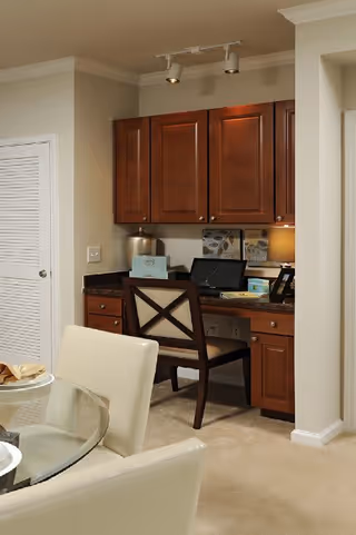 A cozy home office nook with wooden cabinets above and below a granite countertop desk. A wooden chair with a cushioned seat is placed at the desk, which holds a laptop, a tissue box, a lamp, and some decorative items. In the foreground, part of a glass dining table with cream-colored upholstered chairs is visible.
