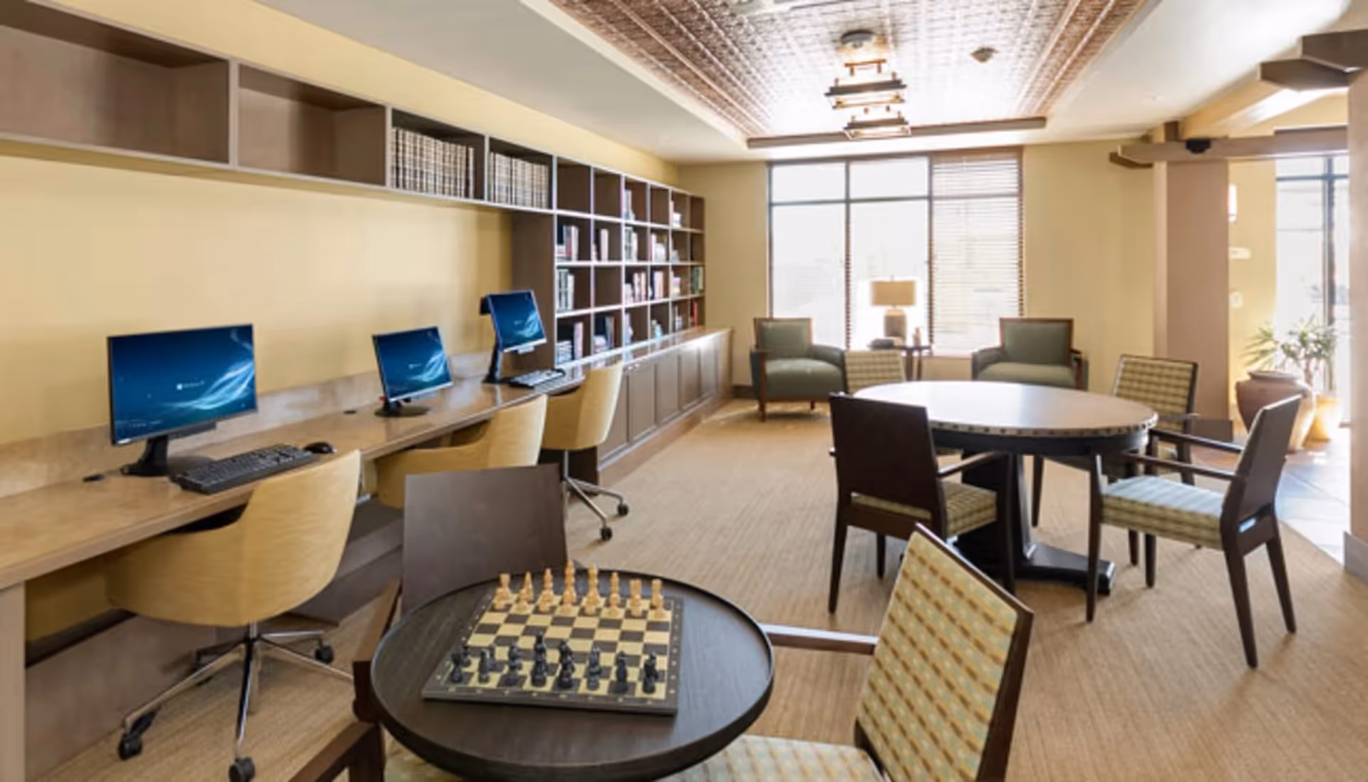 A well-lit room with a row of three computers on a long desk with chairs, a bookshelf filled with books, a round table with four chairs, and another small round table with a chessboard set up. The room has large windows letting in natural light and a decorative ceiling.