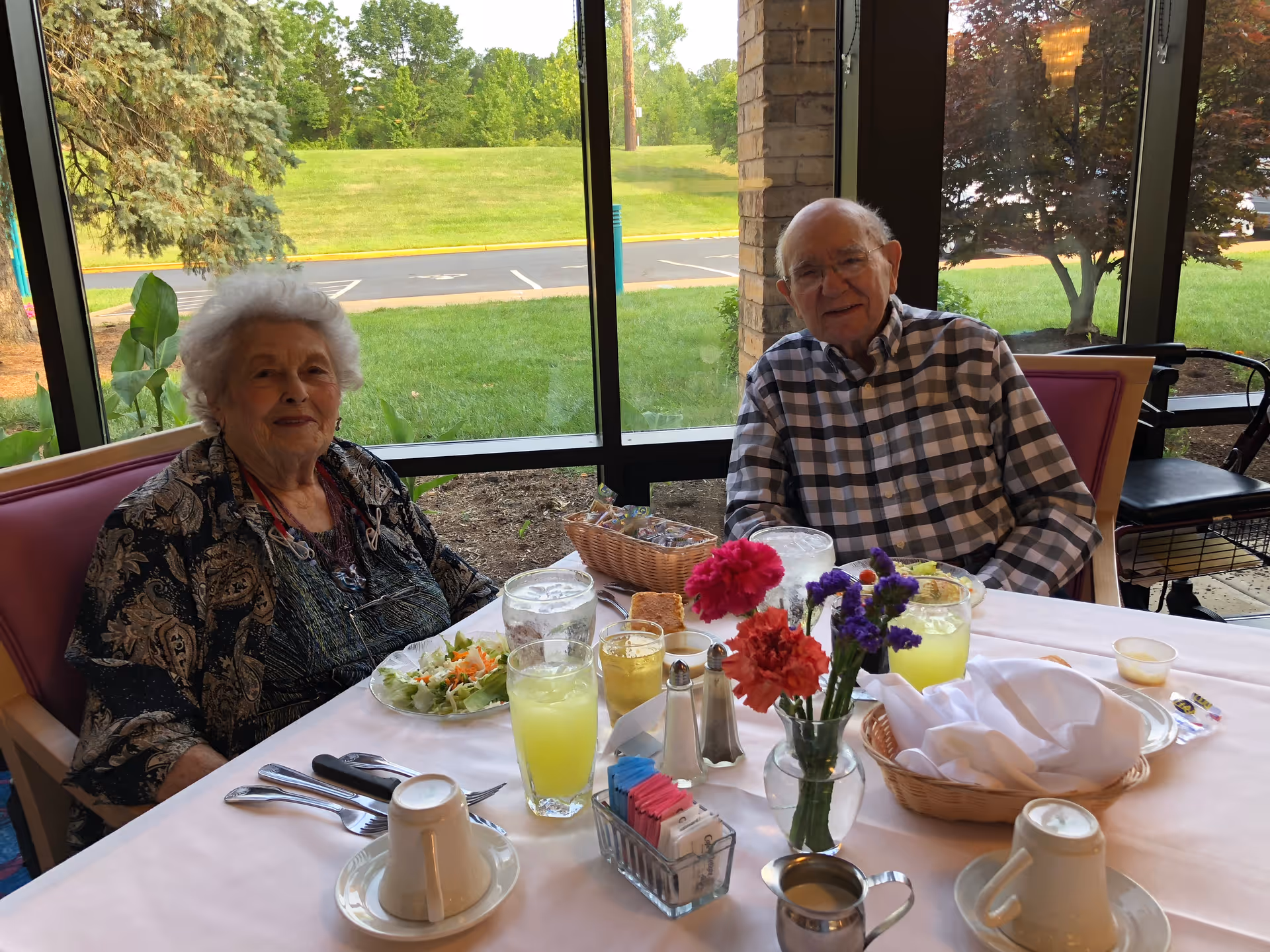 Two elderly people sitting at a dining table inside a room with large windows showing a green outdoor area. The table is set with glasses of lemonade, plates of food, a basket of bread, a small vase with colorful flowers, and various condiments.