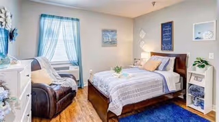 A cozy bedroom with a wooden bed frame, neatly made bed with striped bedding and multiple pillows. There is a dark brown armchair with a light throw blanket near a window with blue curtains. The room has wooden flooring, a white dresser, a small white shelf with decor items, and framed artwork on the walls.